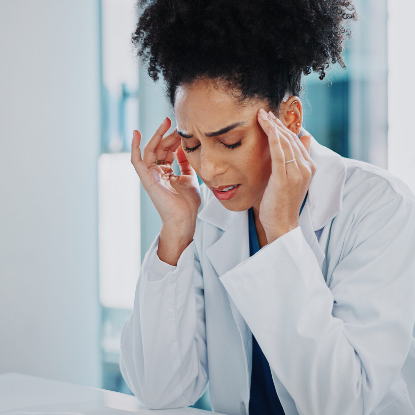 Doctor, woman and headache at hospital with stress, anxiety and burnout at desk with pain. Frustrated, medical and healthcare employee with brain fog, tired and fatigue from clinic issue of surgeon